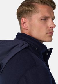 Veste bleu marine avec un col haut et une texture douce. Comprend une section zippée et des accents de boutons, mettant en valeur un design superposé.