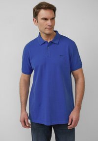 Blauwe polo gemaakt van gestructureerde stof, met een kraag, drie knopen en een klein zwart logo op de linkerborst. Korte mouwen.