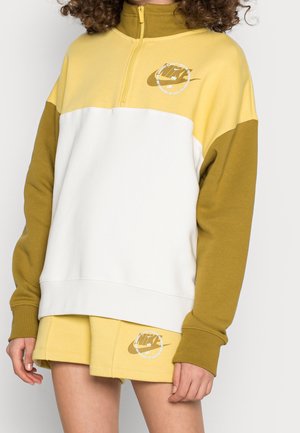Personne portant un sweat-shirt bicolore jaune, blanc et olive avec une fermeture éclair quart de zip et un short jaune assorti avec des logos sur les deux articles.