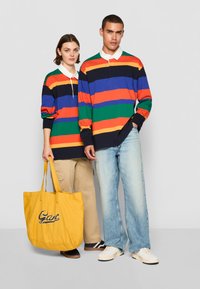 Langærmede poloer med striber i marineblå, orange, blå, grøn og rød; med en hvid krave. En gul tote bag med blå tekst bæres.