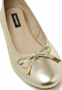 Gold metallic ballet flat with a rounded toe, featuring a small bow detail with gold accents and a smooth, shiny surface.