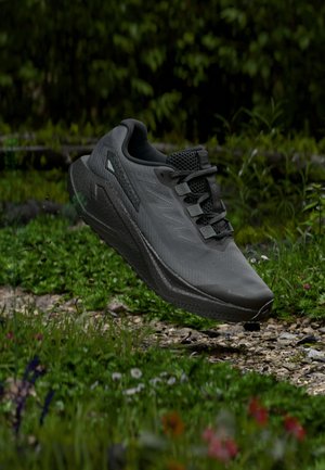 Black trail running shoe with thick sole mid-air above rocky and grassy forest path, surrounded by green plants and wildflowers.