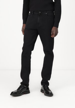 Sorte denimjeans med rett benpassform, med knappelukking og framlommer, brukt med blanke sorte sko.