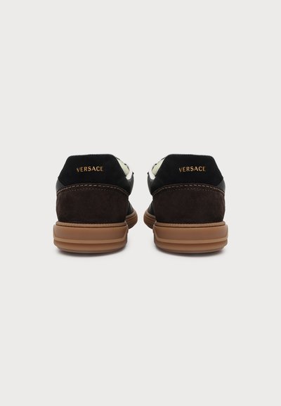 Paire de baskets Versace marron et noires vues de dos, avec une marque Versace dorée sur les talons et des semelles en caoutchouc beige.
