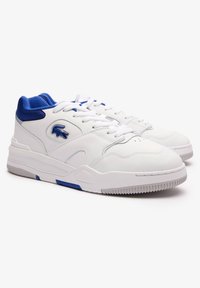 Baltas sneakeru ar ziliem akcentiem, ar gludu ādas virsu, apaļu purngalu, mīkstu apkakli un teksturētu gumijas zoli. Lacoste logo sānos.