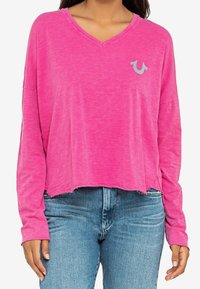 Roze longsleeve V-hals shirt met een relaxte fit en een ruwe zoom, voorzien van een klein blauw logo op de linkerborst. Draag met blauwe jeans.