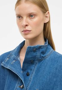 Denim jakna v srednjem modrem odtenku z visokim ovratnikom, vidnimi šivi in kovinskimi gumbi. Tekstura se zdi gladka z nekaj zbledelimi poudarki.