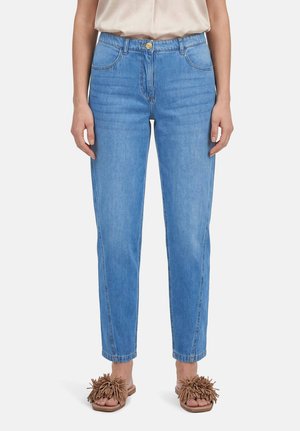 Personne portant un jean droit bleu clair, un haut beige et des sandales marron à franges, debout devant un fond uni.