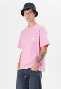 Pink bomulds T-shirt med korte ærmer, der har et hvidt logo på venstre bryst og en afslappet pasform, parret med mørke denimjeans og en sort bøttehat.