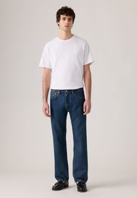 White short-sleeve t-shirt worn with loose-fitting, dark blue denim jeans. Model stands in black shoes against a neutral background.