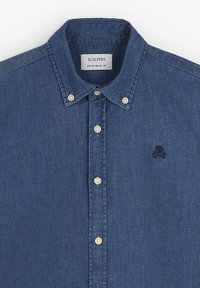 Camicia in denim con colletto a bottoni, bottoni bianchi e un piccolo logo ricamato a scuola e ossa incrociate scuro sul petto.