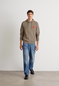 Sudadera con cremallera marrón con un parche de logo rojo, combinada con vaqueros azul claros y zapatos negros, modelo de pie contra un fondo liso.