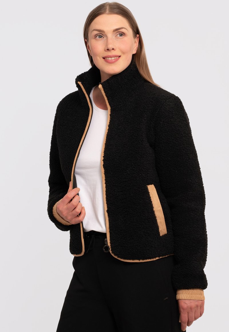 Giacca in fleece nera con dettagli beige, colletto alto e chiusura con zip. La texture è morbida e pelosa, con tasche laterali e polsini a costine.