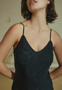 Fekete pántos felső, mély V-nyakkal, finom virágmintás hímzéssel gyöngyökkel, és vékony spagetti pántokkal. Puha, texturált anyag.