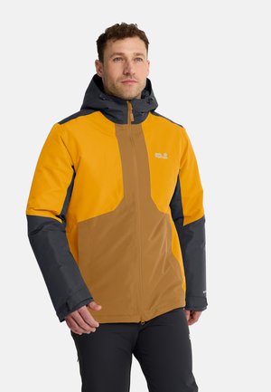 Man draagt een geel, bruin en zwart outdoor jack met kap en zwarte broek, staand tegen een effen lichte achtergrond.