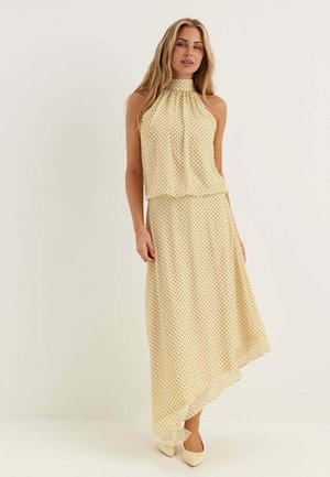 Donna con top beige senza maniche stile halter e gonna lunga asimmetrica abbinata con piccoli pois neri, in piedi davanti a uno sfondo semplice.