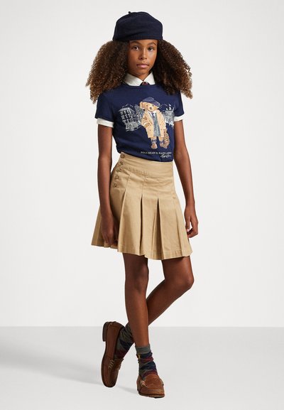 Camiseta de marina con gráfico de oso, falda kaki con pliegues, zapatos marrones y calcetas estampadas; la boina añade un accesorio distintivo.