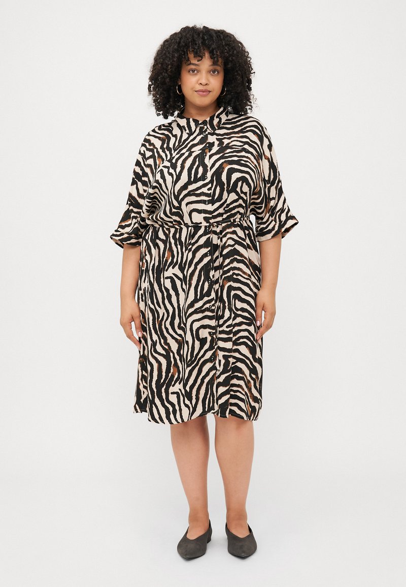 Trumpetas trumpomis rankovėmis, pagamintas iš smėlio spalvos audinio su juodais ir rudais zebro raštais, surištas ties juosmeniu su raišteliu ir dekoruotas mygtukais.