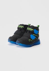 Sapatilhas de crianças Skechers pretas e azuis com detalhes verdes, correias de velcro e solas azuis translúcidas sobre um fundo branco.