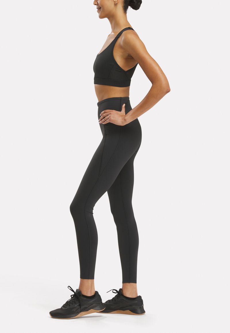 Sujeción deportiva negra y leggings de cintura alta, ambos en una tela suave y elástica. Zapatillas deportivas negras con suela texturizada. Vista lateral.
