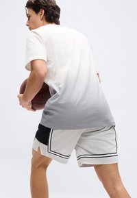 Νεαρός άνδρας που φοράει γκρί πουκάμισο με ομπρέ εφέ και λευκά σορτς μπάσκετ, κρατώντας μια μπάλα του μπάσκετ, έτοιμος να κάνει κίνηση.