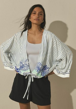 Woman wearing a loose, striped kimono jacket with blue coral prints over a white top and black shorts, standing against a beige wall.