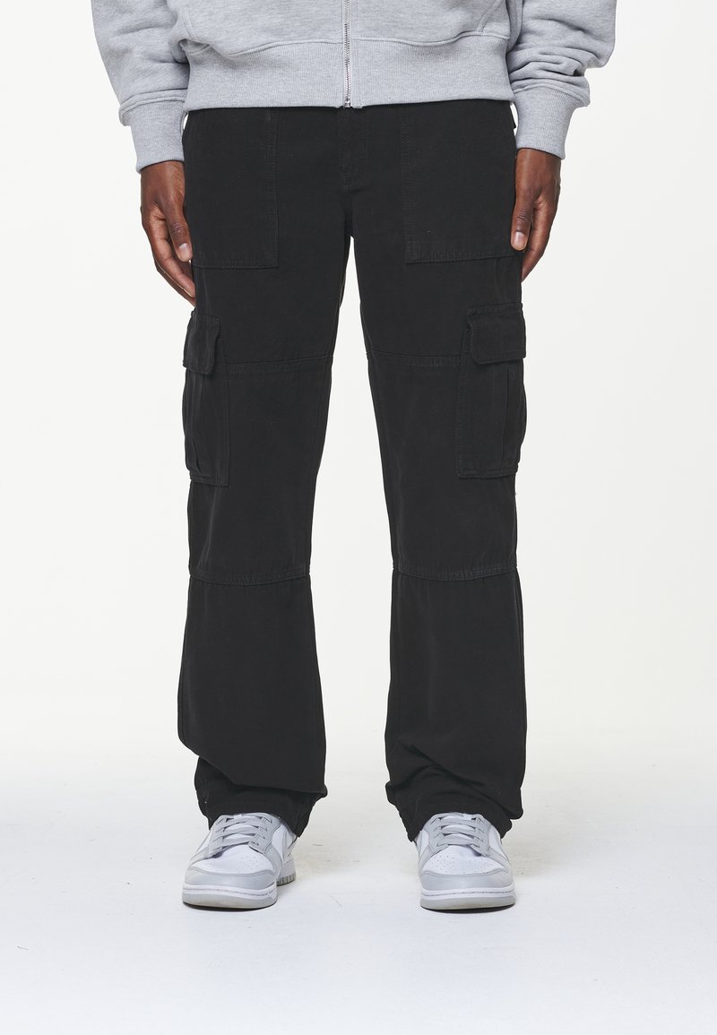 Homme portant un pantalon cargo noir et des baskets blanches, debout devant un fond clair uni avec les mains détendues le long du corps.