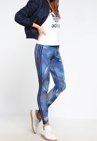 Blå leggings med virvlande abstrakta mönster, vita ränder längs sidorna, kombinerat med en croppad vit t-shirt och en marinblå bomberjacka.