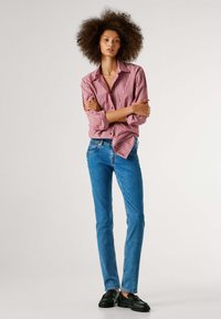 Rød- og hvitstripet knappeskjorte med lange ermer, ikke stukket inn, kombinert med blå smale jeans og svarte loafers på en modell med krøllete hår.