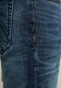 Vaqueros de mezclilla en un lavado azul oscuro con un acabado texturizado. Presentan una etiqueta negra distintiva con "CHASIN'" en la costura lateral.