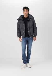Sort pufferjakke med hætte, udstyret med en frontlynlås og trykknapper. Båret over en sort skjorte og parret med blå jeans og hvide sneakers.