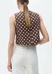 Hnědý top bez rukávů s puntíky a zkráceným střihem, doplněný o světlé džíny s kapsami na zadní straně.