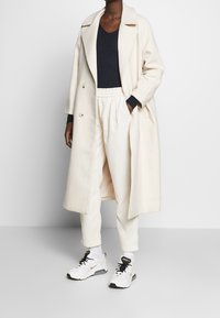 Dwurzędowy płaszcz w kolorze kremowym z mieszanki wełny, zestawiony z prążkowanymi, jasnymi spodniami oraz białymi butami sportowymi z czarnymi akcentami.
