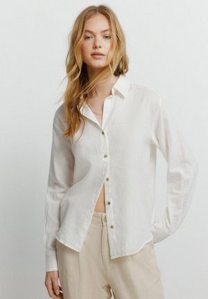 Młoda kobieta z długimi blond włosami, ubrana w białą koszulę na guziki i beżowe spodnie, stojąca z jedną ręką na biodrze na jednolitym tle.