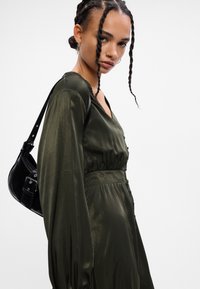 Σκούρο πράσινο σατέν φόρεμα με μακριά μανίκια, ντεκολτέ σε σχήμα V και μαζεμένη μέση. Το μοντέλο κρατάει μια μικρή μαύρη τσάντα χεριού με λουρί στον ώμο.