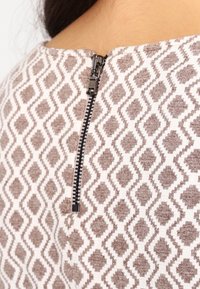 Tissu à motifs avec un design de diamants beige et marron. Présente une fermeture éclair noire au dos du col pour la fermeture.