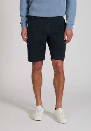 Hombre vestido con pantalones cortos azul marino, suéter de manga larga azul claro y zapatillas blancas, de pie frente a un fondo liso.