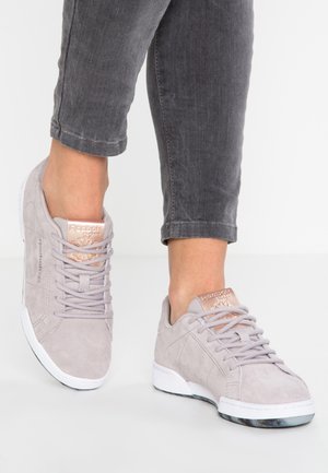 Person iført lysegrå Reebok Classic sneakers med rosaguld logo detaljer og mørkegrå cropped jeans mod en hvid baggrund.