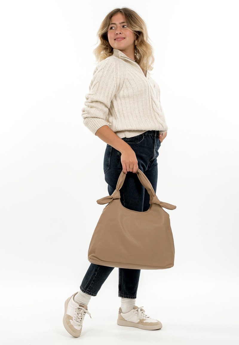 Beige leather handbag with tied handles, spacious design. Model wearing cream sweater, dark jeans, and white sneakers stands against a white background.