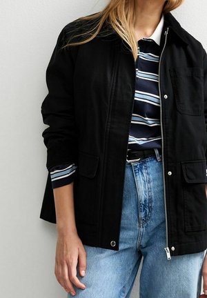 Personne portant une veste noire sur une chemise rayée bleu marine et blanche avec un jean bleu clair, debout devant un fond uni clair.