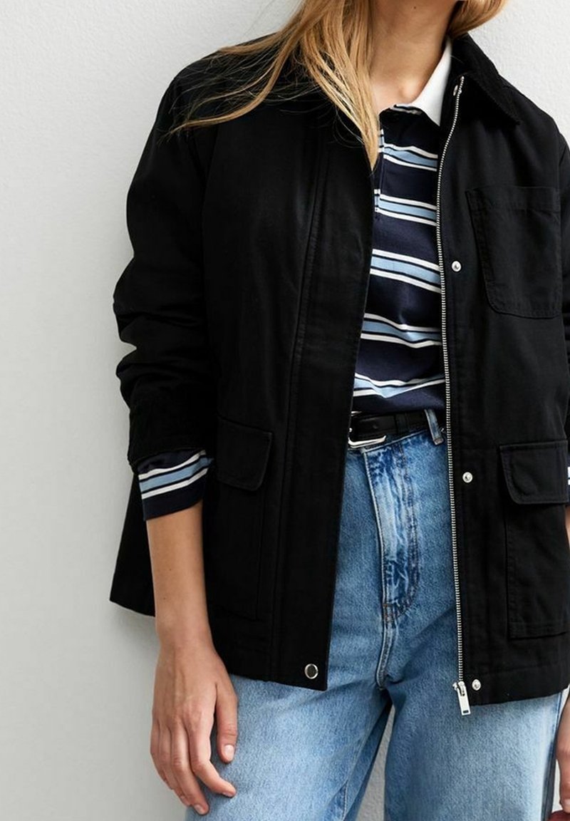 Personne portant une veste noire sur une chemise rayée bleu marine et blanche avec un jean bleu clair, debout devant un fond uni clair.