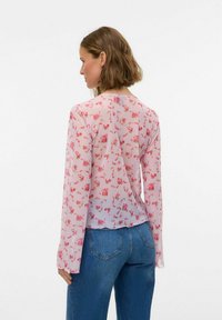 Blouse légère et transparente de couleur rose avec un petit motif floral, manches longues, encolure ronde et ourlet légèrement arrondi. Portée avec un jean bleu.