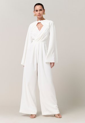 Kvinde iført en hvid vidbenet jumpsuit med vidt udstående lange ærmer, knyttet halsudskæring og guldringeøreringe, stående foran en neutral baggrund.
