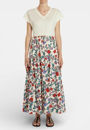 Donna che indossa un top crema a maniche corte con scollatura a V e una gonna lunga a strati con motivo floreale rosso, viola e verde, in piedi con sandali.