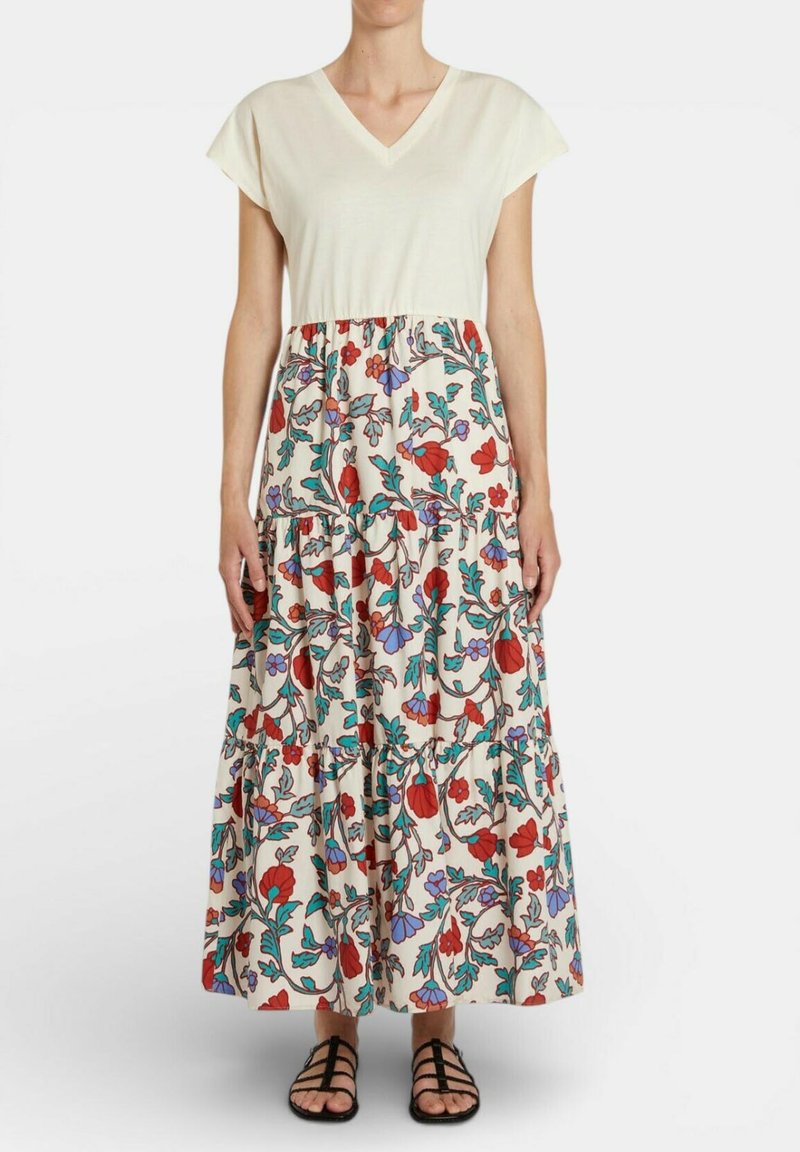 Donna che indossa un top crema a maniche corte con scollatura a V e una gonna lunga a strati con motivo floreale rosso, viola e verde, in piedi con sandali.