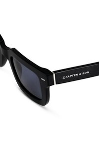 Gafas de sol negras con montura rectangular, lentes oscuros y detalles en plata. Presenta el logo "KAPTEN & SON" en la patilla.