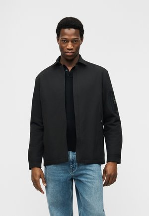 Młody mężczyzna w czarnej kurtce zapinanej na zamek, na czarnej koszulce polo i jasnoniebieskich dżinsach, stojący na tle jednolitego białego tła.