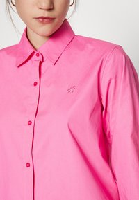 Camicia button-up rosa realizzata in tessuto morbido, con colletto classico, bottoni coordinati e un logo ricamato discreto sul petto.