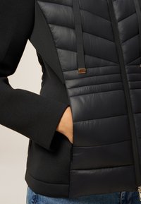 Person, der eine schwarze gesteppte Weste mit Reißverschluss und Kordelzügen trägt, die Hand in die Seitentasche gesteckt hat, kombiniert mit einem schwarzen Langarm-Shirt und blauen Jeans.
