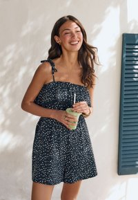 Tânără cu păr lung și castaniu, purtând o rochie alb-neagră cu buline, zâmbind și ținând un smoothie verde într-un pahar transparent.